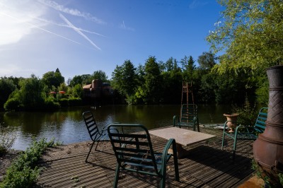 Miniature Location salle Gif-sur-Yvette (Essonne) - Villa Utopia #36 Terrasse en bois avec des chaises autour d'une table, donnant sur un étang entouré de végétation et d'arbres.