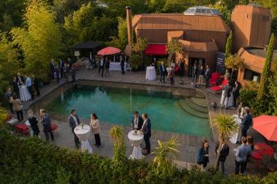 Piscine extérieure entourée de végétation, avec terrasse aménagée de chaises et parasols rouges sous un ciel bleu.