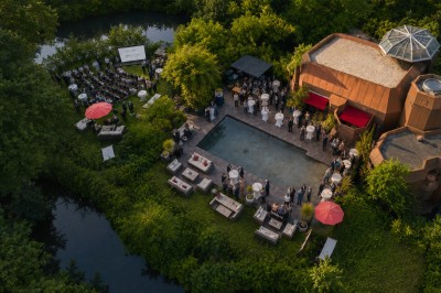 Piscine extérieure entourée de végétation, avec terrasse aménagée de chaises et parasols rouges sous un ciel bleu.