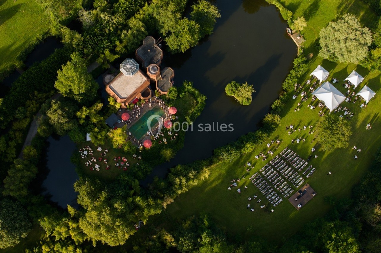 Vue aérienne d'un domaine avec un bâtiment entouré d'eau, des tentes blanches et des chaises disposées en extérieur.