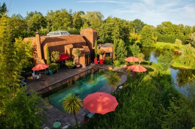 Piscine extérieure entourée de végétation, avec terrasse aménagée de chaises et parasols rouges sous un ciel bleu.