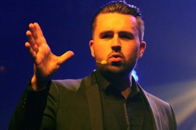Un homme barbu pose devant un fond vert avec des cartes de trèfle flottant et des faisceaux lumineux.