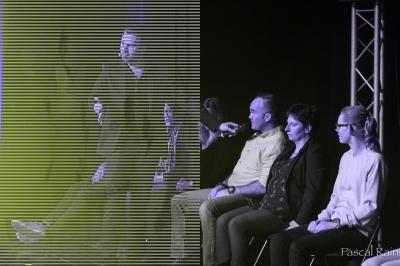 Un homme barbu pose devant un fond vert avec des cartes de trèfle flottant et des faisceaux lumineux.