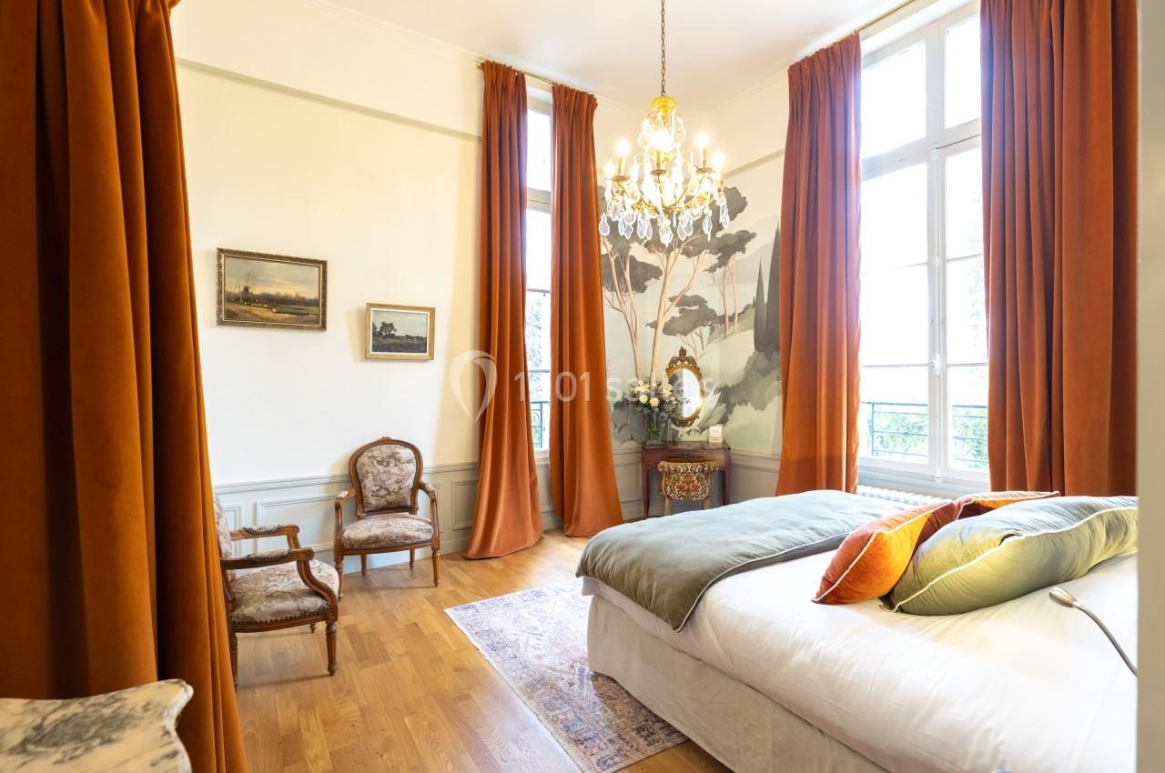 Chambre lumineuse avec lit double, parquet, rideaux orange, lustre et grandes fenêtres donnant sur l'extérieur.