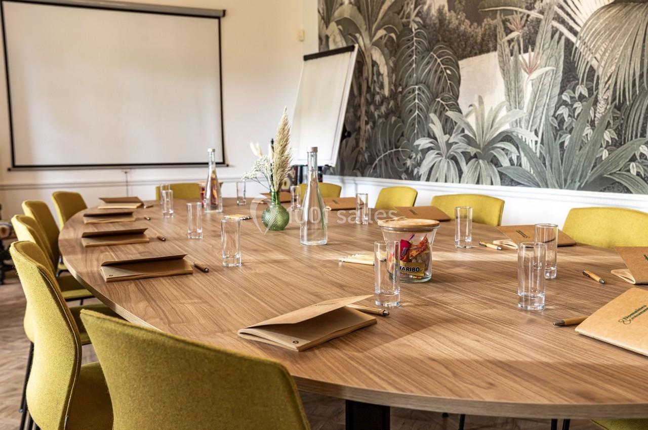Salle de réunion avec table ovale en bois, chaises jaunes, carnets, verres et décor mural végétal en arrière-plan.