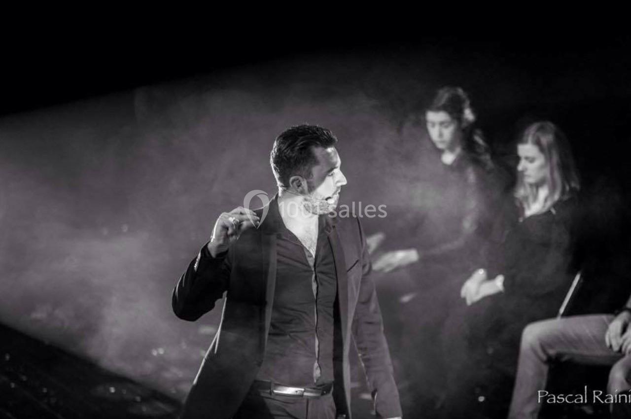 Un homme en costume parle sur scène dans une ambiance sombre et enfumée, avec des spectateurs en arrière-plan.
