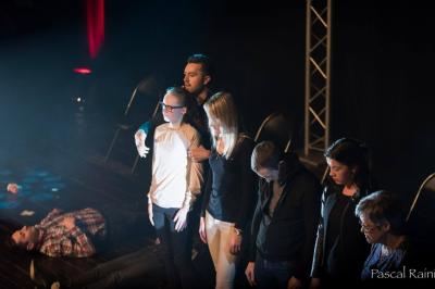 Un homme sur scène interagit avec quatre personnes assises, éclairées par une lumière contrastée.