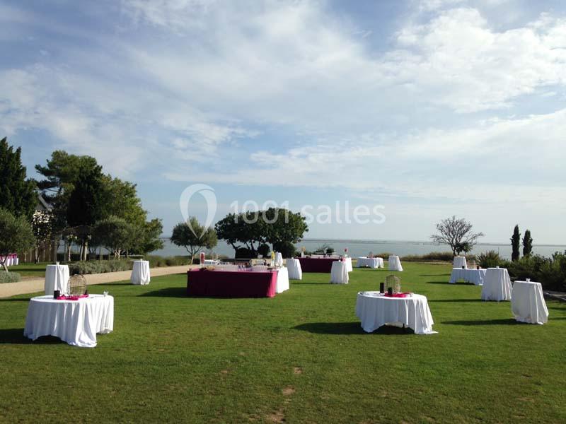 Tables dressées avec nappes blanches sur une pelouse, entourées d'arbres, avec vue sur un paysage côtier.