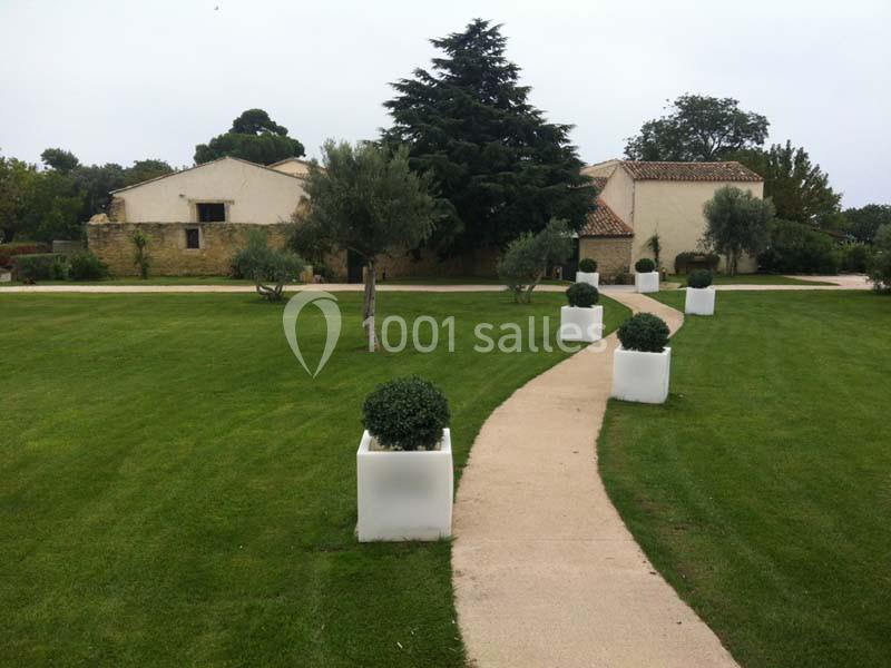 Allée pavée bordée de buis en pots blancs, menant à des bâtiments en pierre entourés de pelouses et d'arbres.