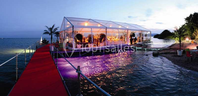 Tente transparente éclairée installée sur une plateforme au bord de l'eau, avec un tapis rouge menant à l'entrée.