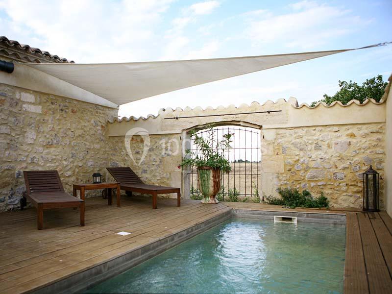 Petite piscine extérieure entourée de murs en pierre, avec terrasse en bois, transats et voile d'ombrage.