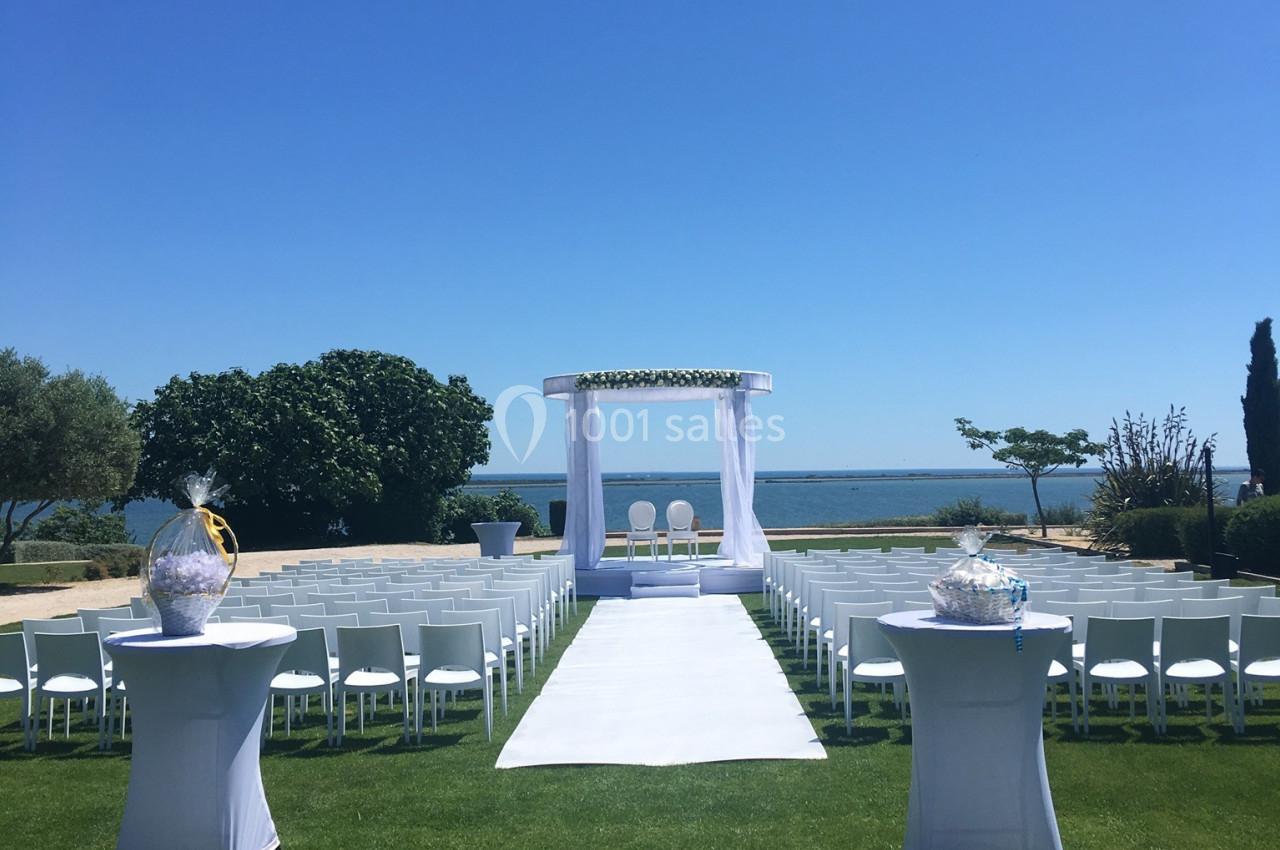 Allée centrale menant à une arche de mariage blanche, entourée de chaises, avec vue sur la mer et un ciel dégagé.