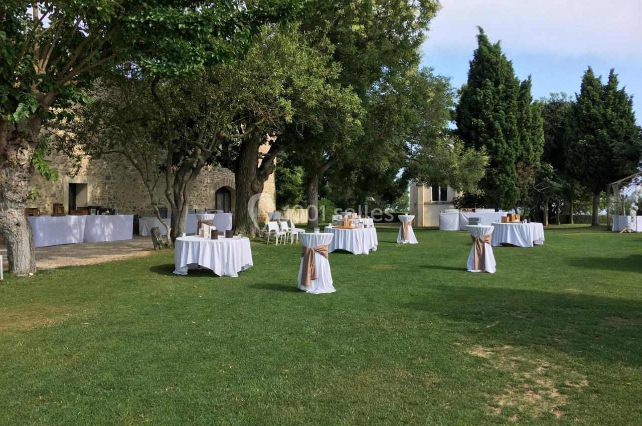 Tables rondes dressées avec nappes blanches et décorations beige, disposées sur une pelouse entourée d'arbres.