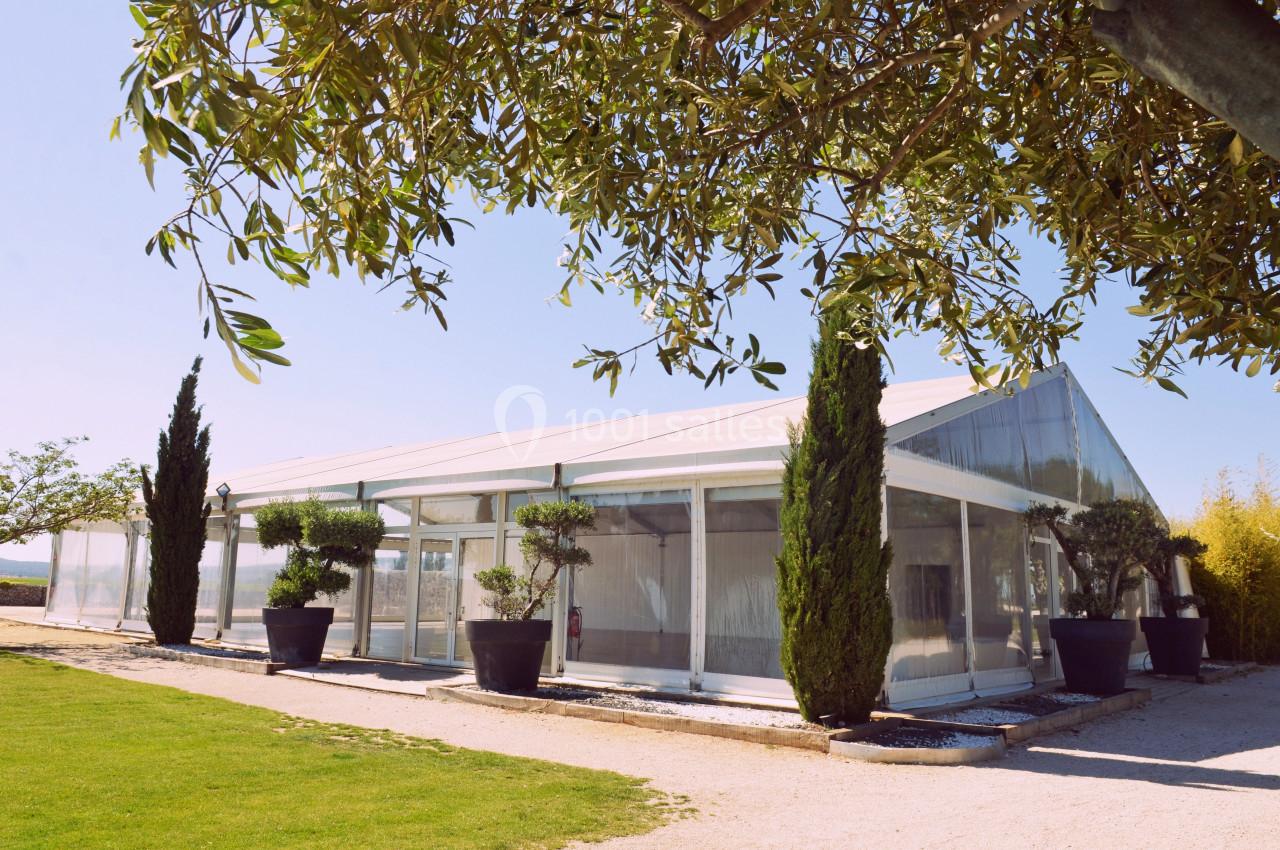 Structure en verre et métal entourée de plantes et d'arbres, située sur un chemin bordé de pelouse.