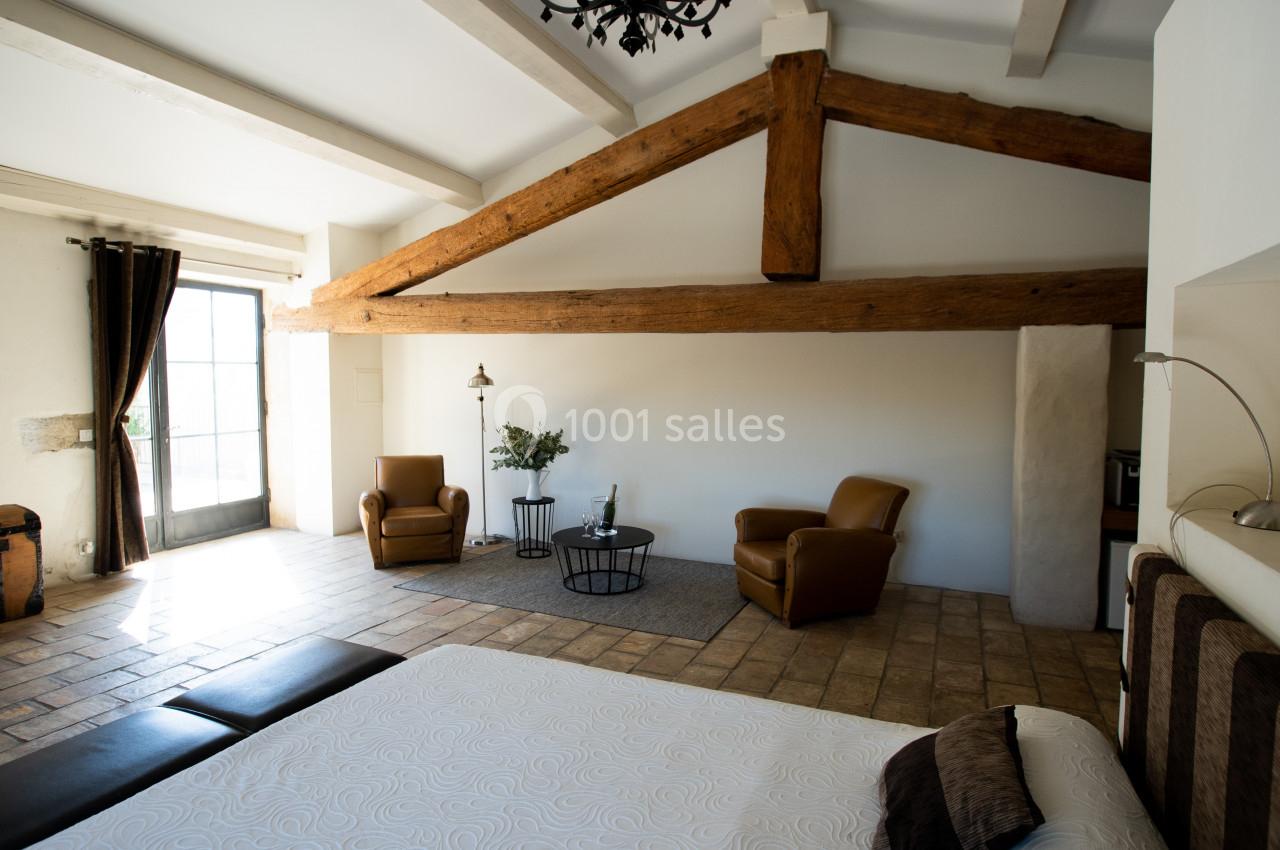 Chambre lumineuse avec poutres apparentes, deux fauteuils, un lit, une malle en bois et une grande porte-fenêtre.