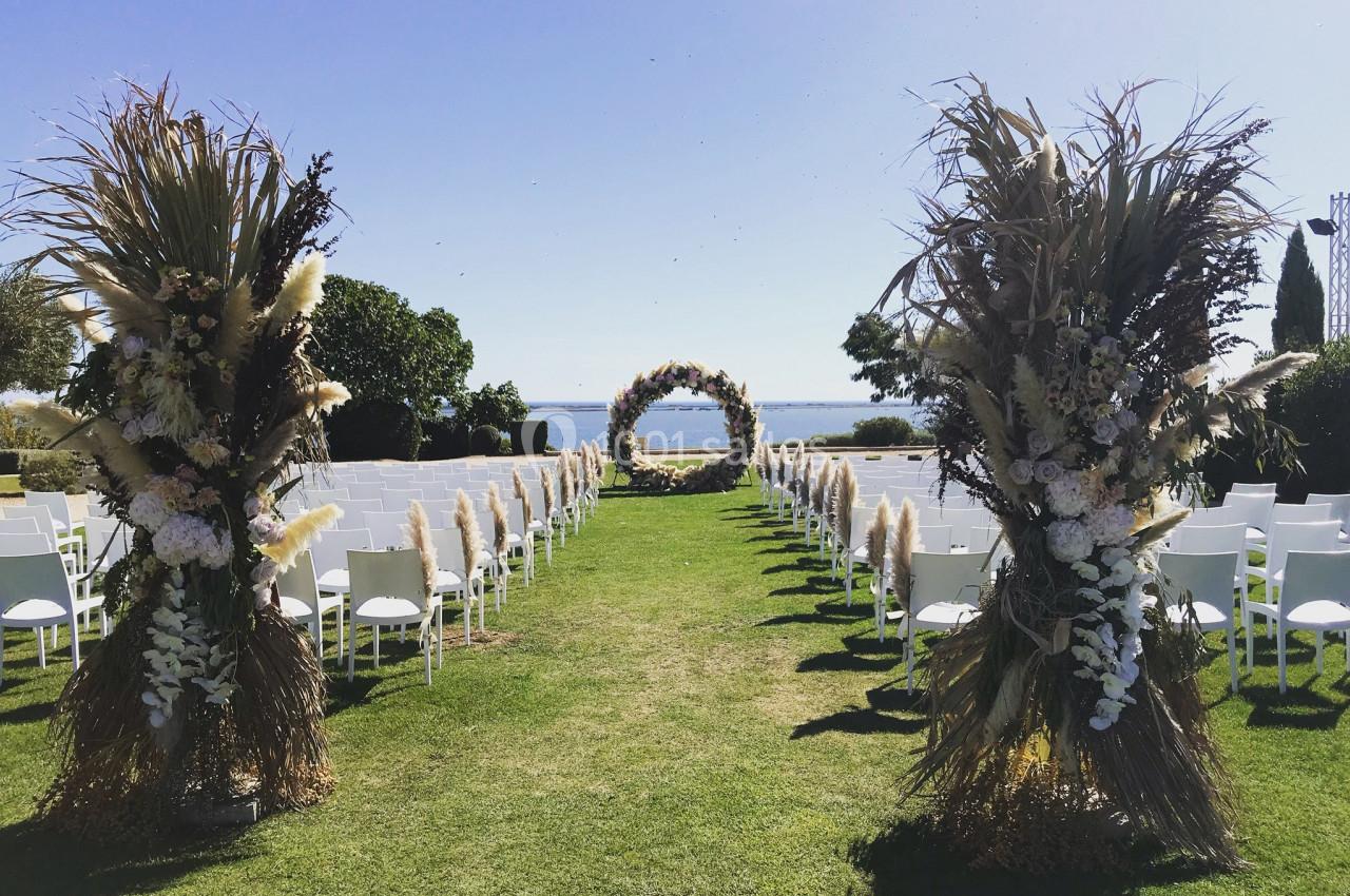 Allée centrale décorée de fleurs séchées menant à une arche florale sur une pelouse, avec vue sur la mer.