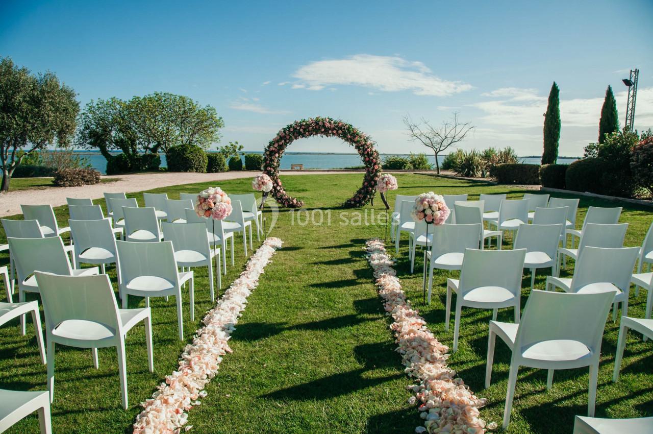 Chaises blanches alignées sur une pelouse face à une arche florale, décorée pour une cérémonie en extérieur.