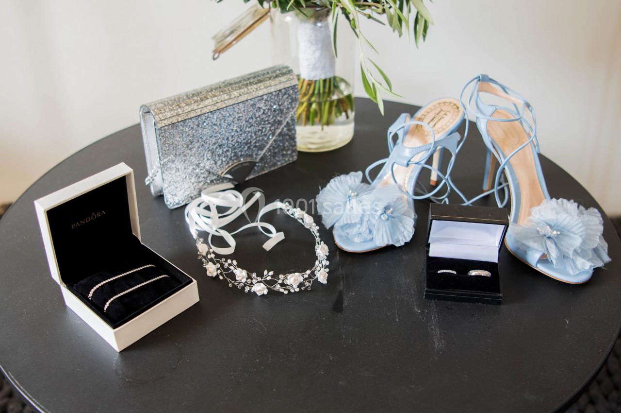 Accessoires de mariage sur une table : chaussures bleues, sac pailleté, bijoux et alliances dans des écrins.