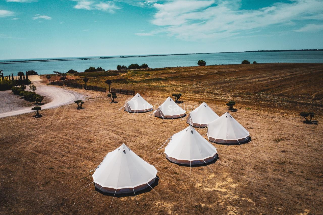 Tentes blanches installées sur un terrain sec près d'un champ et d'un plan d'eau sous un ciel partiellement nuageux.