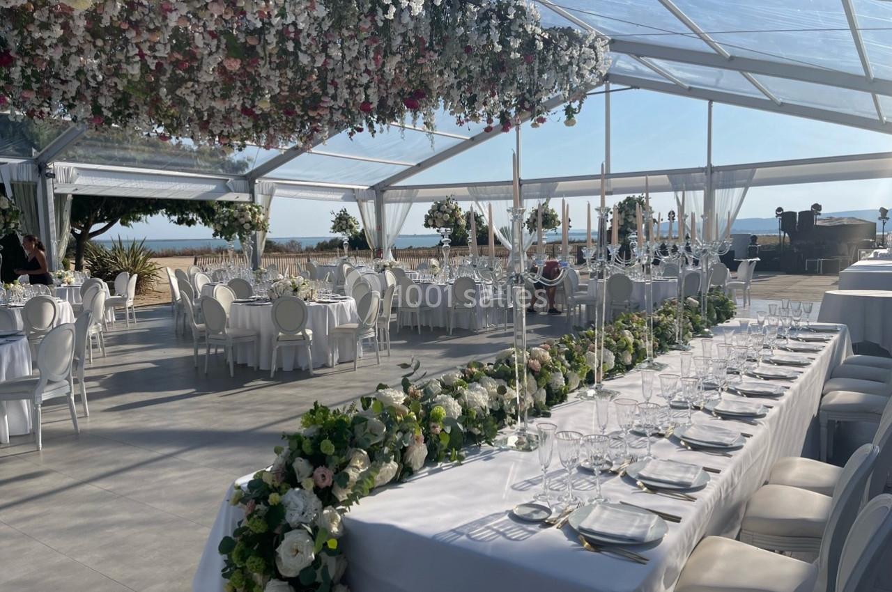 Salle de réception en plein air sous une verrière, décorée de fleurs suspendues et de tables élégantes avec vue sur la mer.