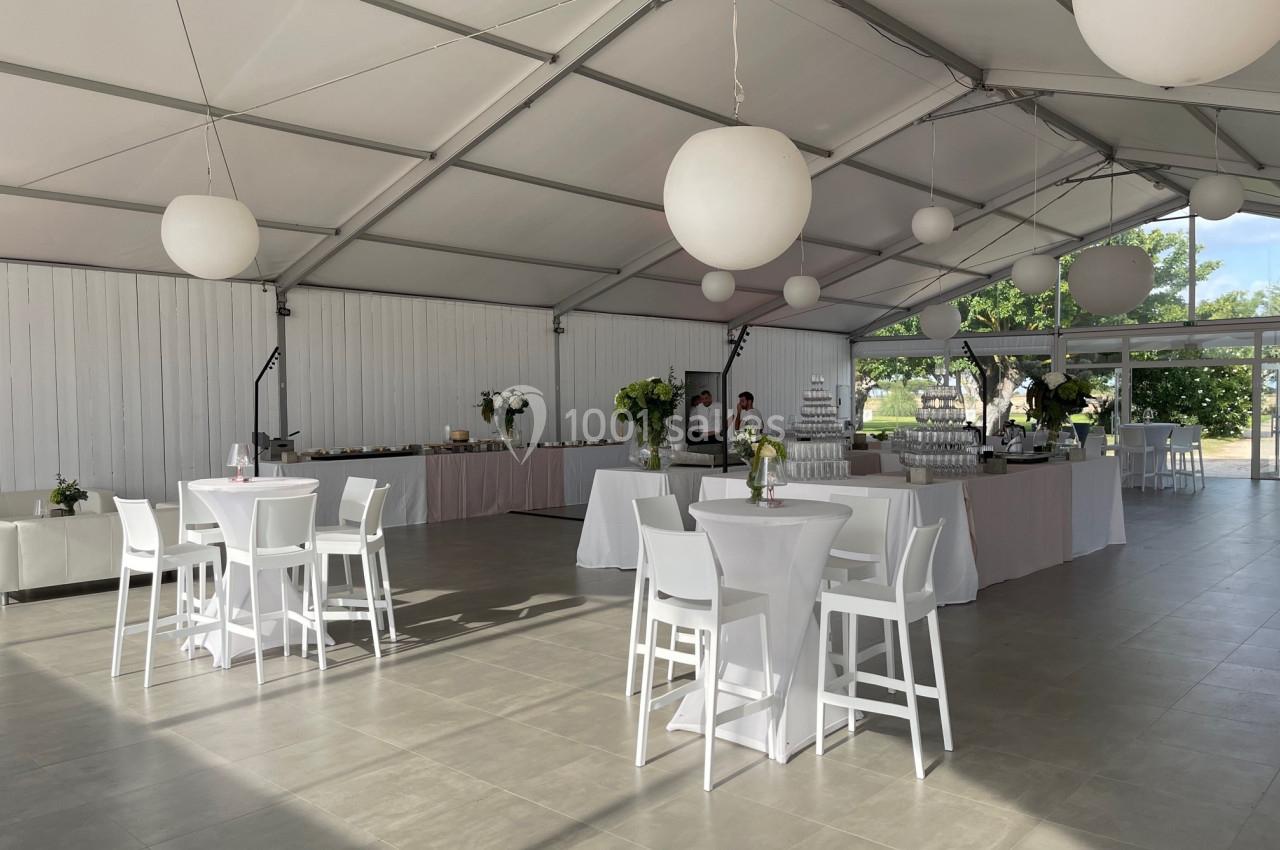 Salle de réception lumineuse avec tables hautes blanches, décor floral et buffet en arrière-plan.