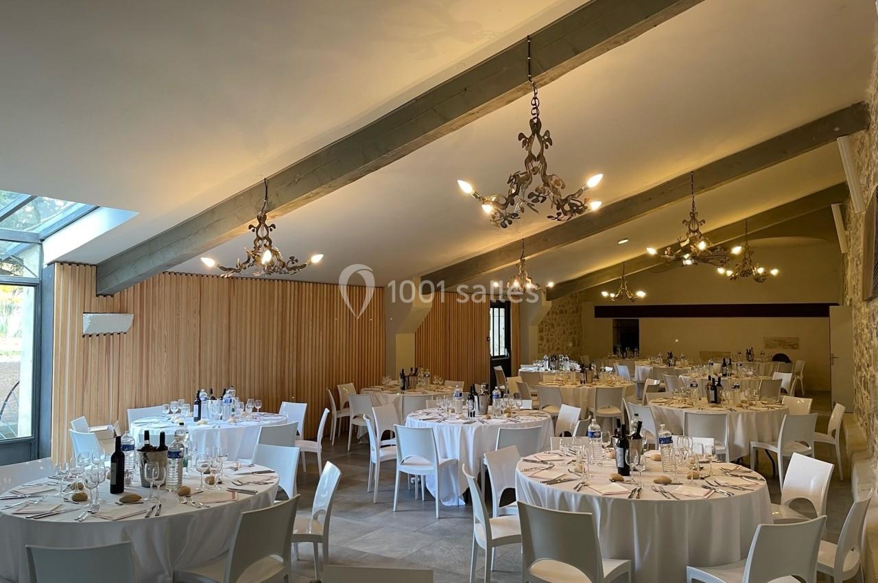 Salle de réception lumineuse avec tables rondes dressées, nappes blanches et chaises modernes sous un plafond avec poutres…