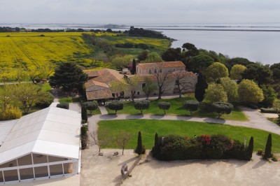 Vue aérienne d'un domaine rural avec bâtiments en pierre, arbres et vue sur une étendue d'eau calme en arrière-plan.