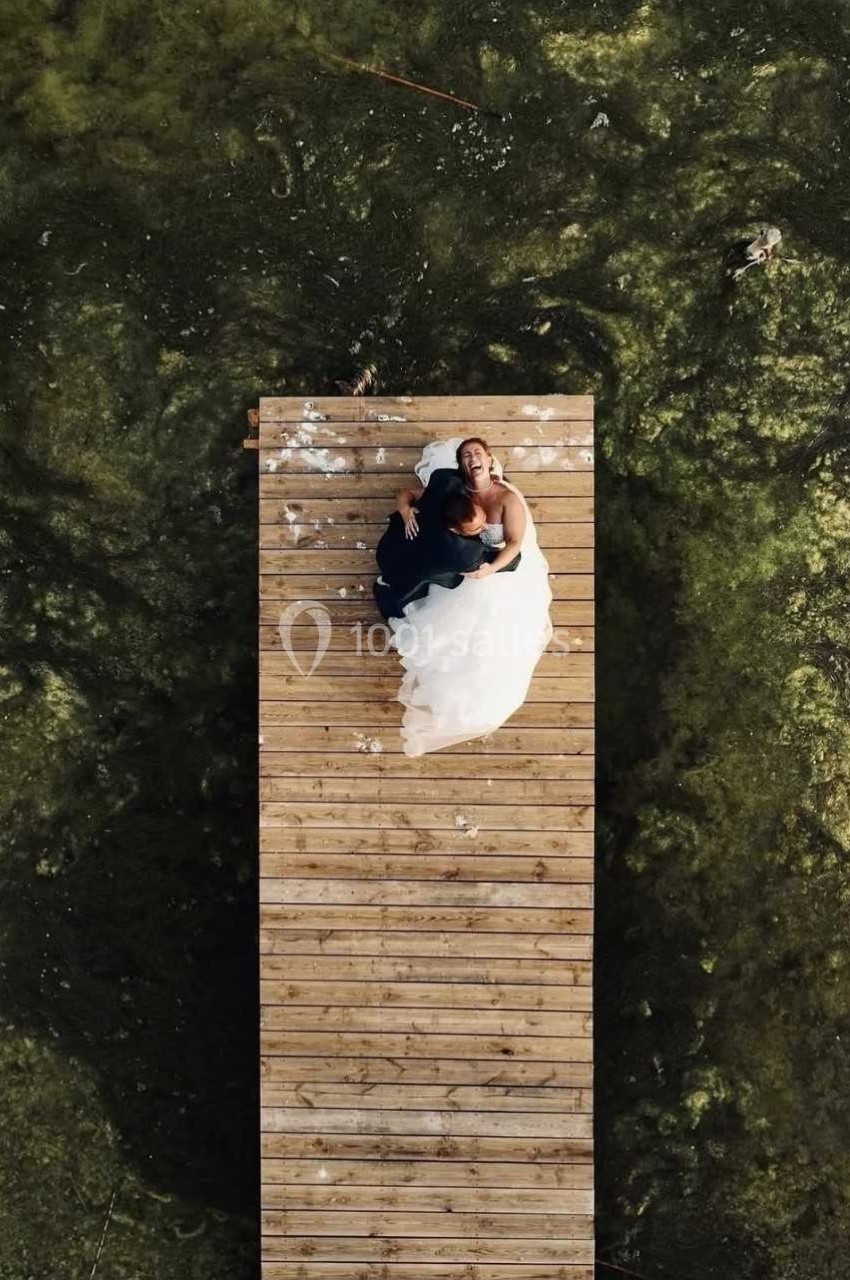 Un couple en tenue de mariage assis sur un ponton en bois au-dessus d'une eau verdâtre vue d'en haut.