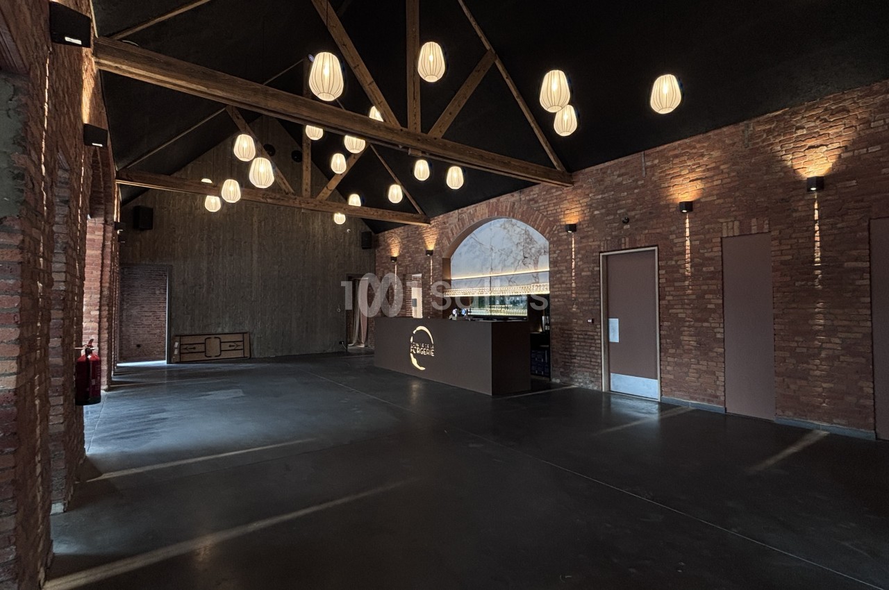 Grande salle vide au plafond en charpente apparente et aux suspensions en forme de lanterne, murs en briques rouges, avec un bar en arc de marbre au fond.