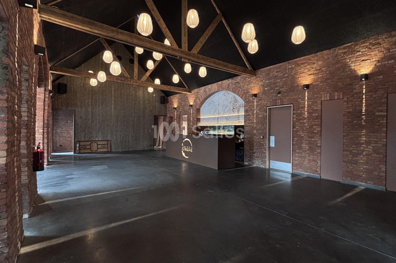 Grande salle vide au sol en béton ciré, murs en briques apparentes, poutres en bois au plafond et suspensions en papier éclairant l'espace.