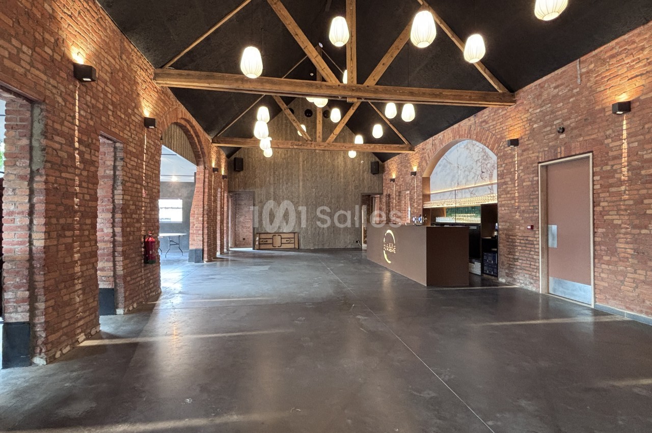 Grande salle vide au sol en béton ciré, murs en briques apparentes, charpente en bois sous plafond sombre et suspensions blanches, comptoir de bar au fond à droite.