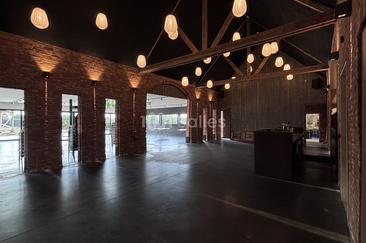 Grande salle vide aux murs de briques apparentes, plafond en charpente bois, suspensions en forme de lanterne, sol en béton ciré et bar au fond à droite.