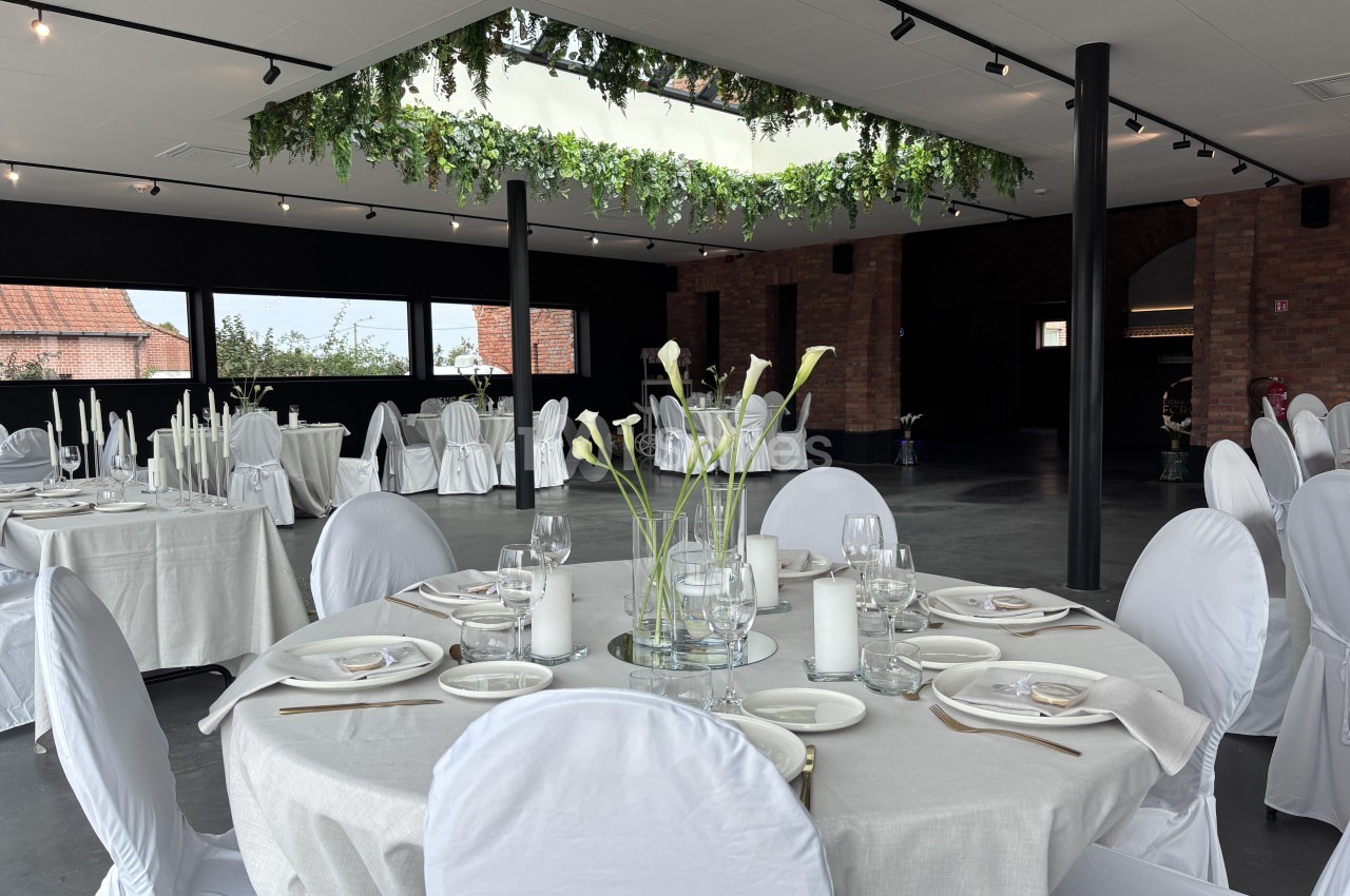 Grande salle aux murs sombres et murs en briques, tables rondes dressées de nappes claires et chaises housses blanches, guirlande de verdure suspendue sous une verrière.