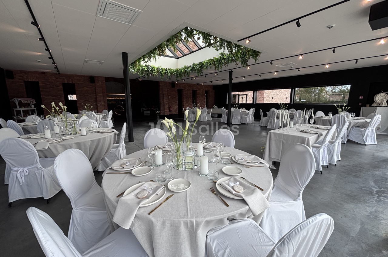 Grande salle aux murs de briques et au sol en béton, dressée de tables rondes nappées de blanc avec chaises housses blanches, sous une verrière ornée de guirlandes végétales. Let me check length: roughly 165 characters, too long. Need ≤125. Shorter