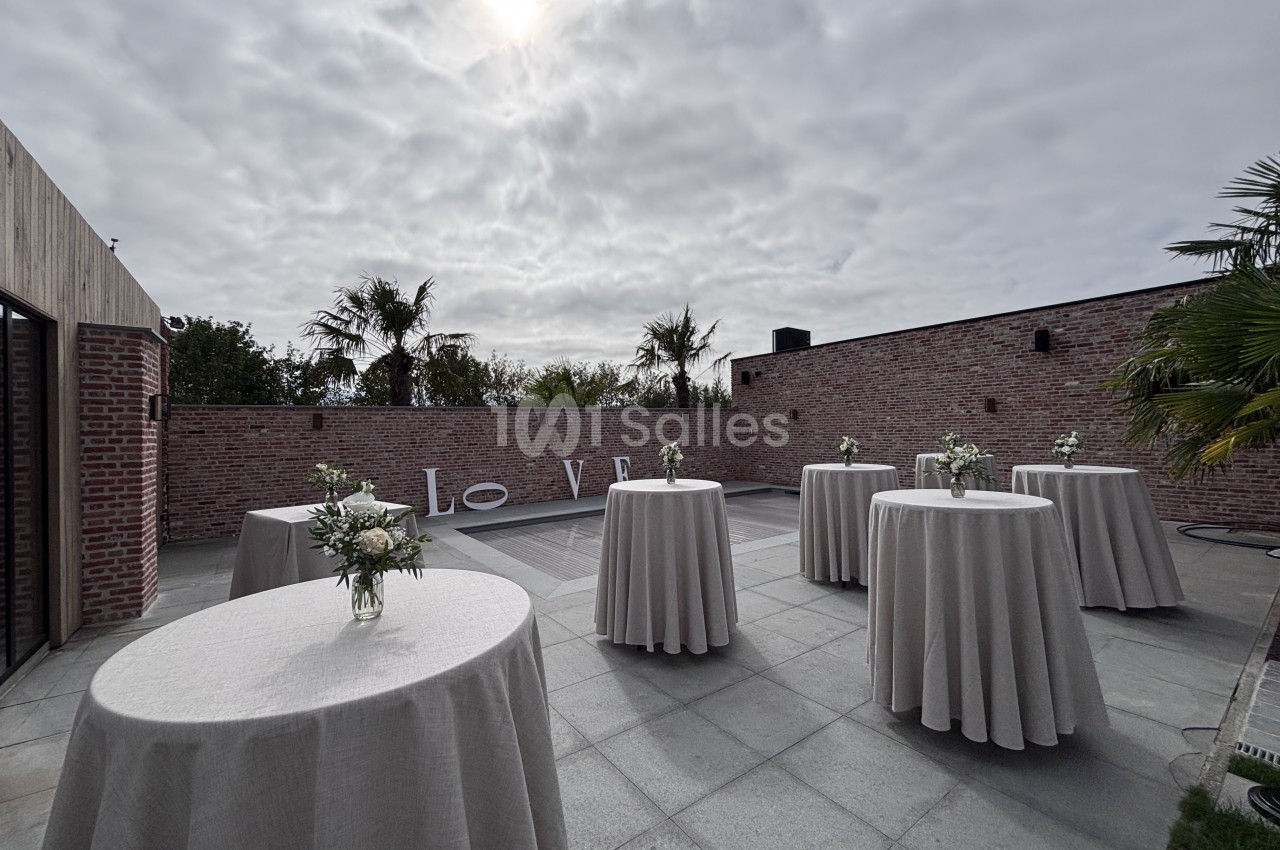 Terrasse extérieure dallée avec plusieurs tables hautes rondes nappées de gris et fleuries, face à un mur en briques et des palmiers.