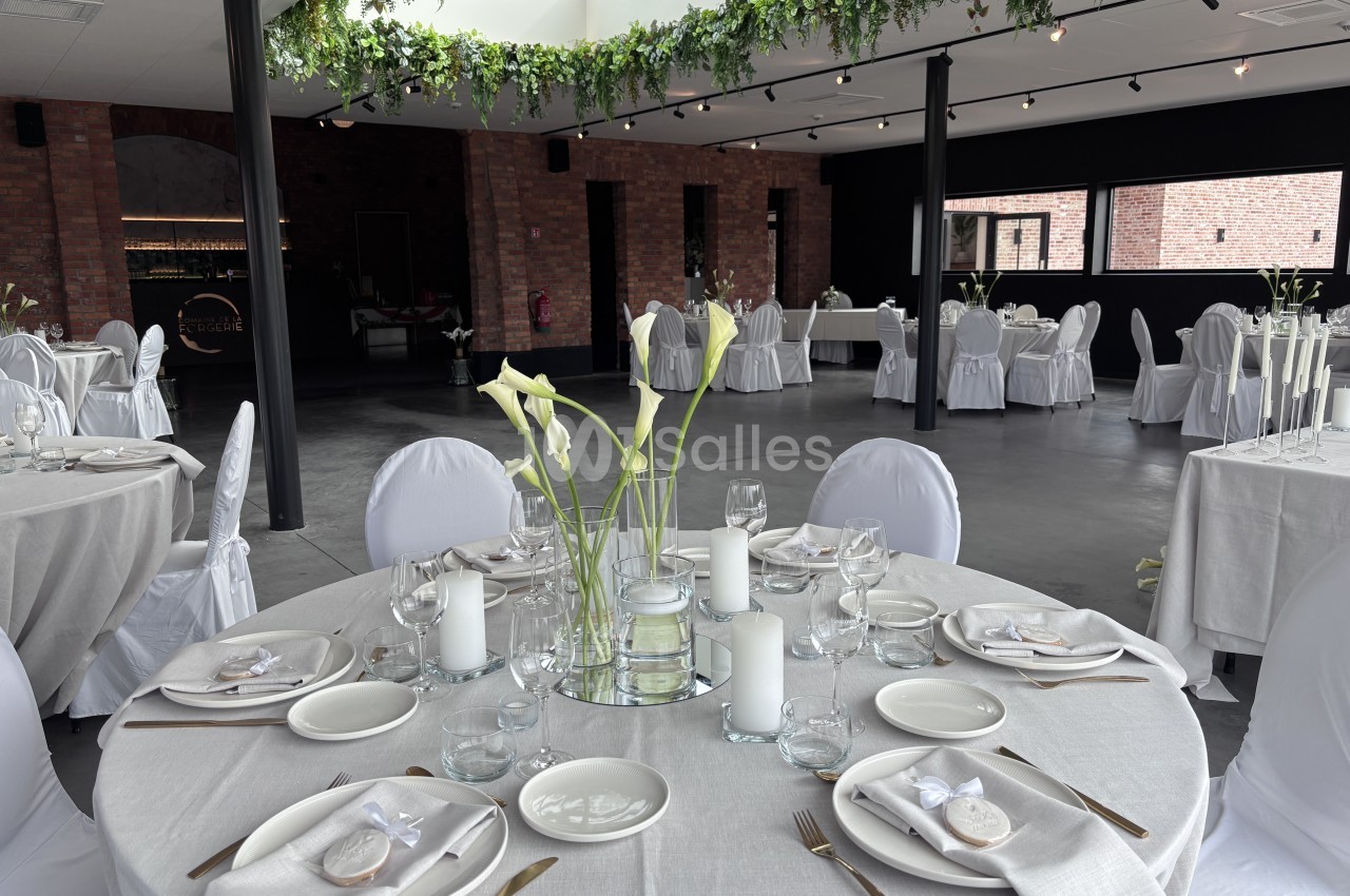 Salle de réception aux murs de briques, tables rondes dressées de nappes grises, serviettes, bougies et bouquets de callas blancs, chaises housées de blanc.