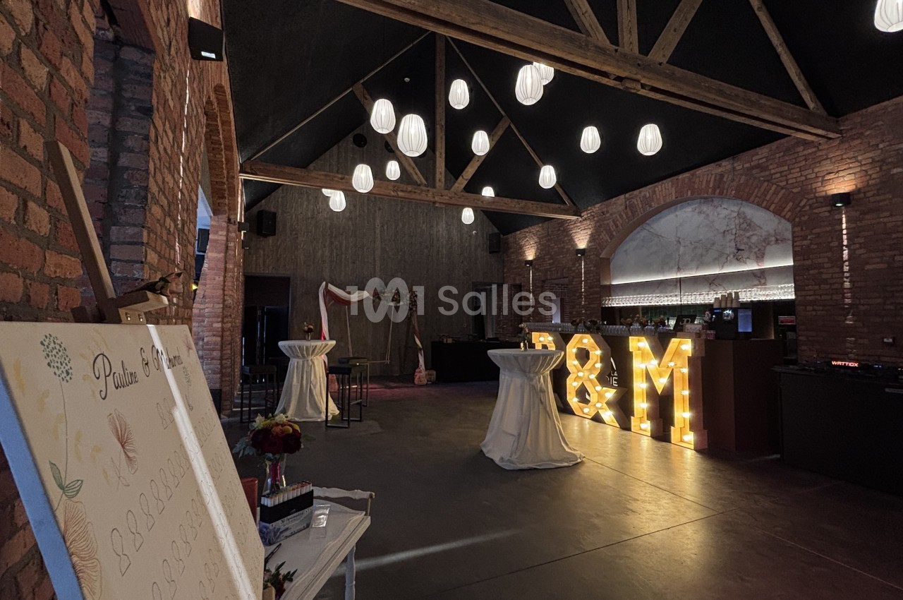 Salle aux murs de briques et poutres apparentes, avec suspensions lumineuses et tables hautes nappées de blanc.