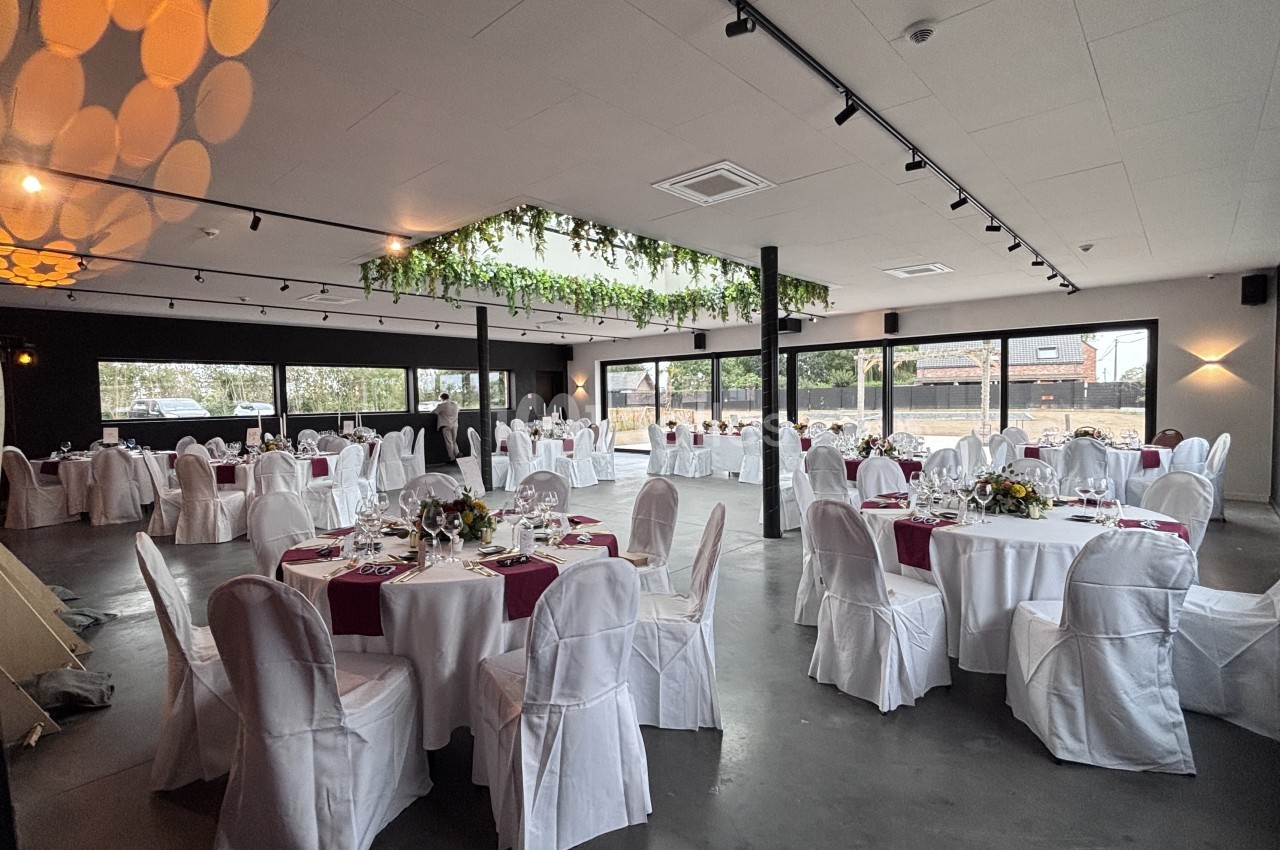 Grande salle de réception aux murs clairs, avec tables rondes nappées de blanc, bordeaux et fleurs jaunes, chaises housses blanches et grandes baies vitrées.