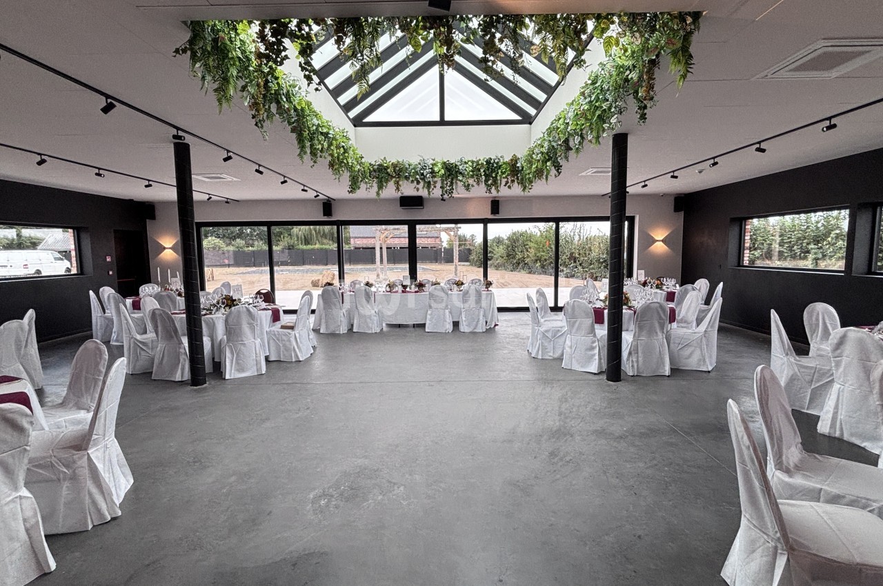 Grande salle de réception aux murs sombres, éclairée par une verrière ornée de guirlandes végétales, avec tables rondes dressées de nappes blanches et chaises habillées de housses blanches.