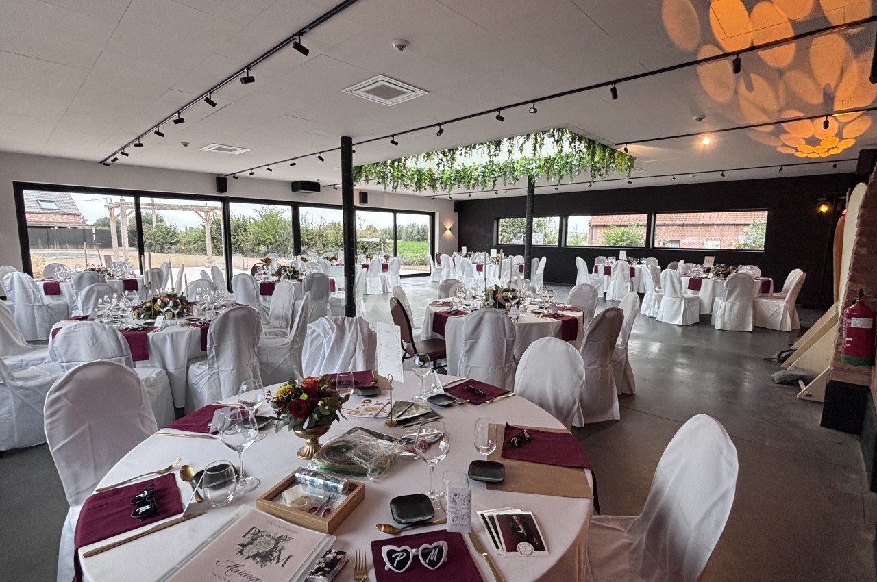 Grande salle de réception aux murs sombres, dressée de tables rondes nappées de blanc avec housses de chaises blanches, serviettes bordeaux et centres de fleurs, éclairée par des projecteurs et de grandes baies vitrées.