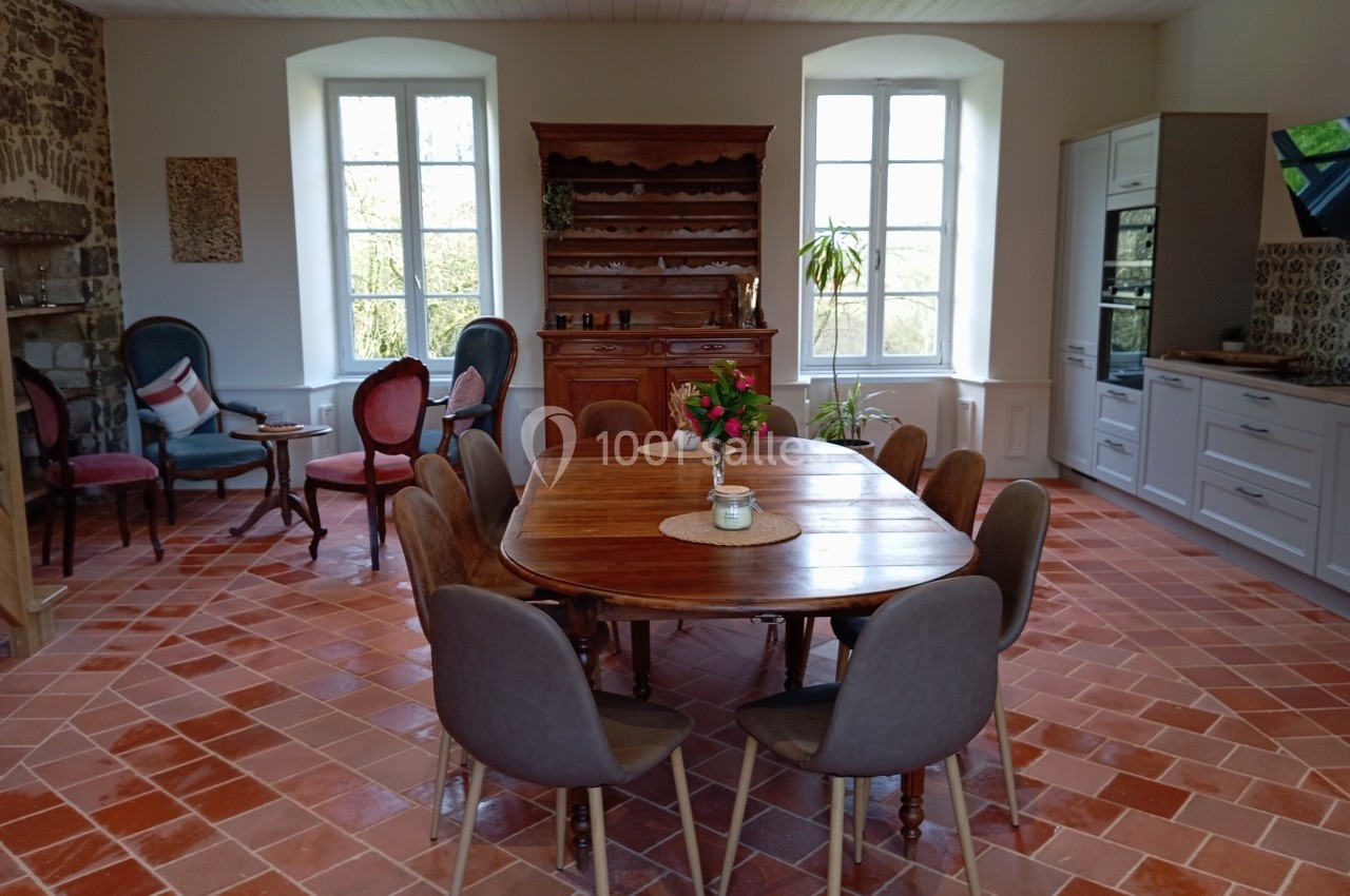 Salle à manger lumineuse avec table en bois entourée de chaises, sol en terre cuite et cuisine équipée en arrière-plan.