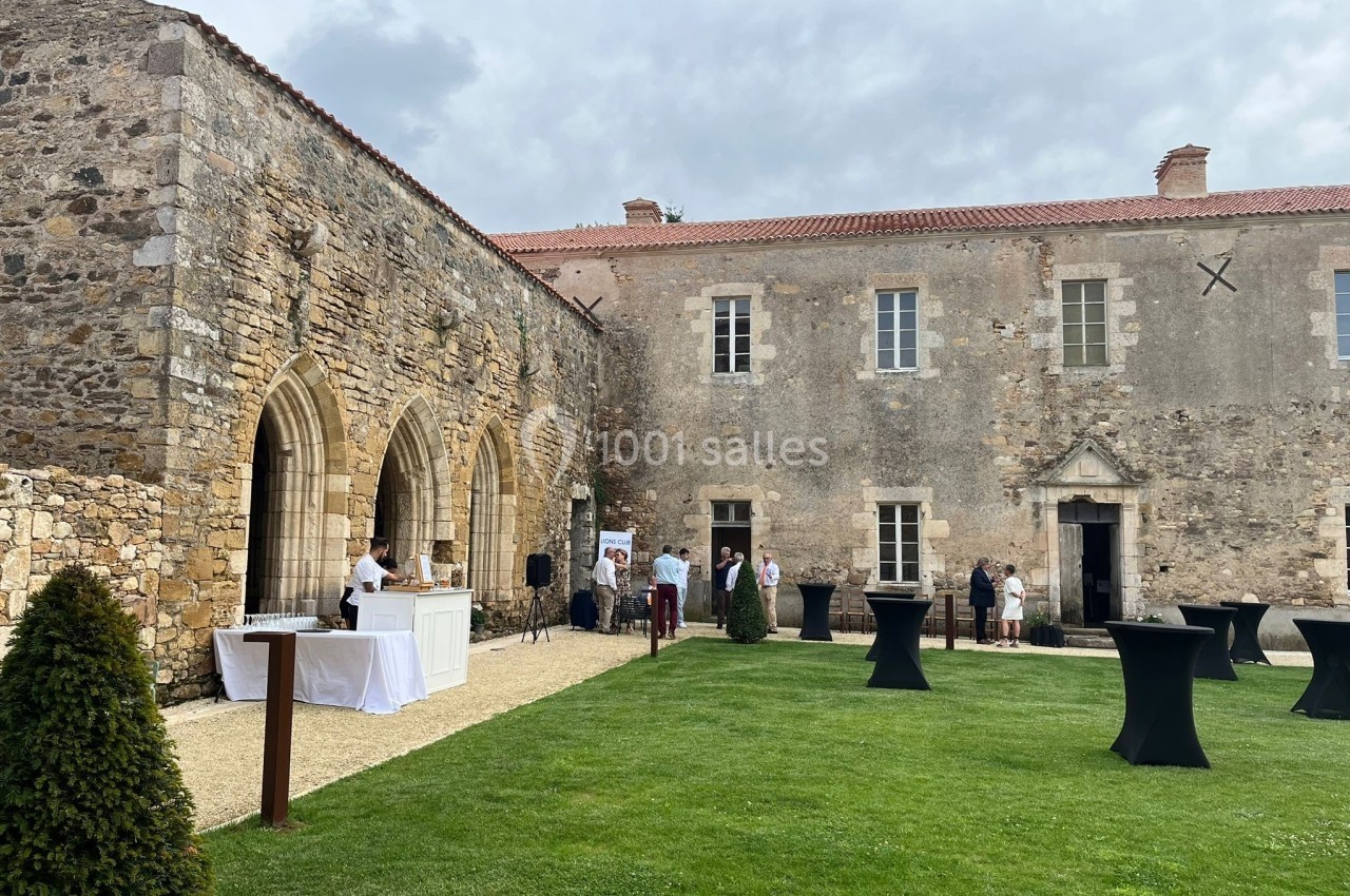 Cour d’un bâtiment en pierre ancienne avec pelouse, tables hautes noires et un stand de service en extérieur.