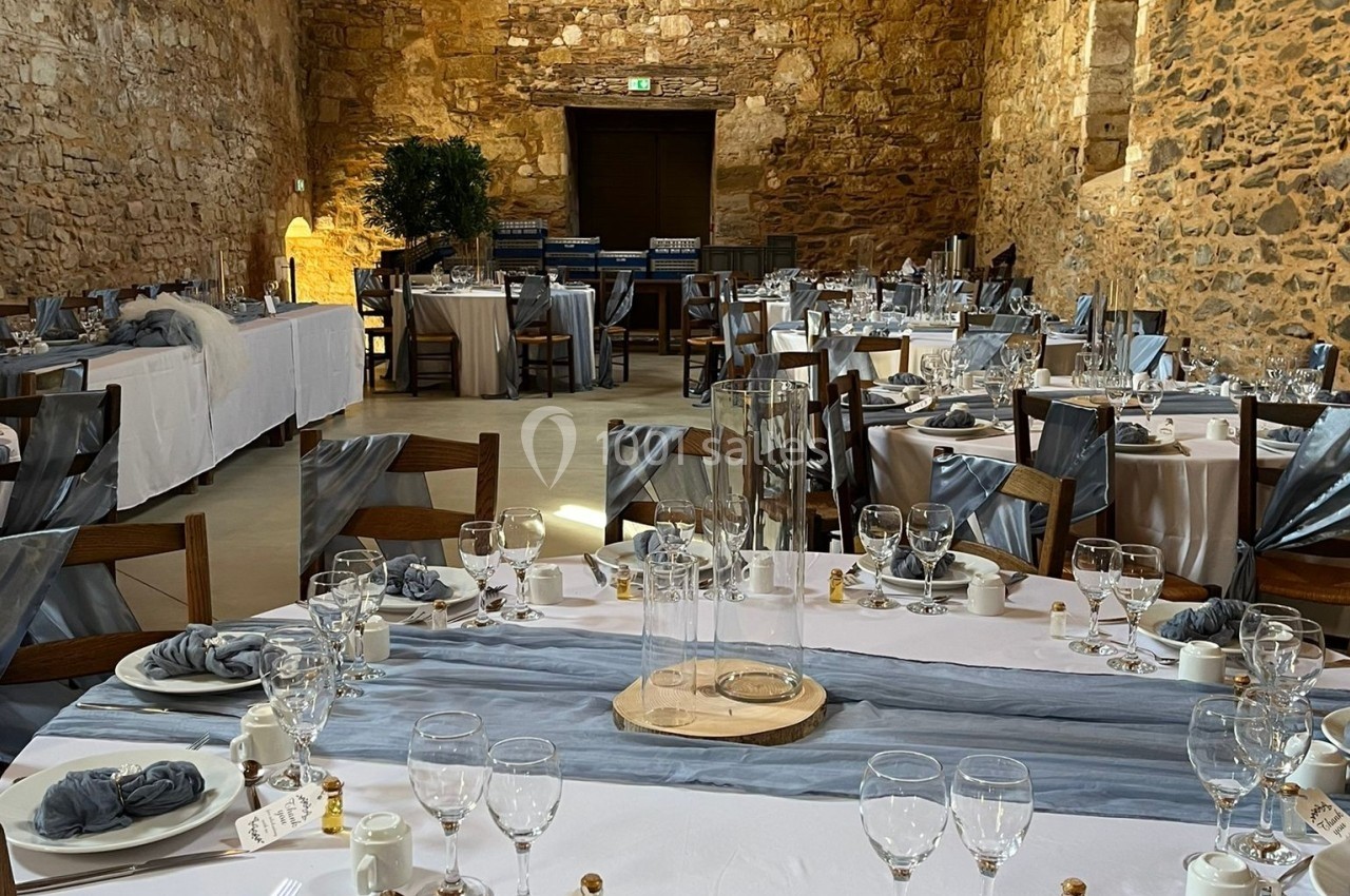 Salle en pierre ancienne aménagée pour un repas, avec tables dressées de nappes blanches et décorations élégantes.