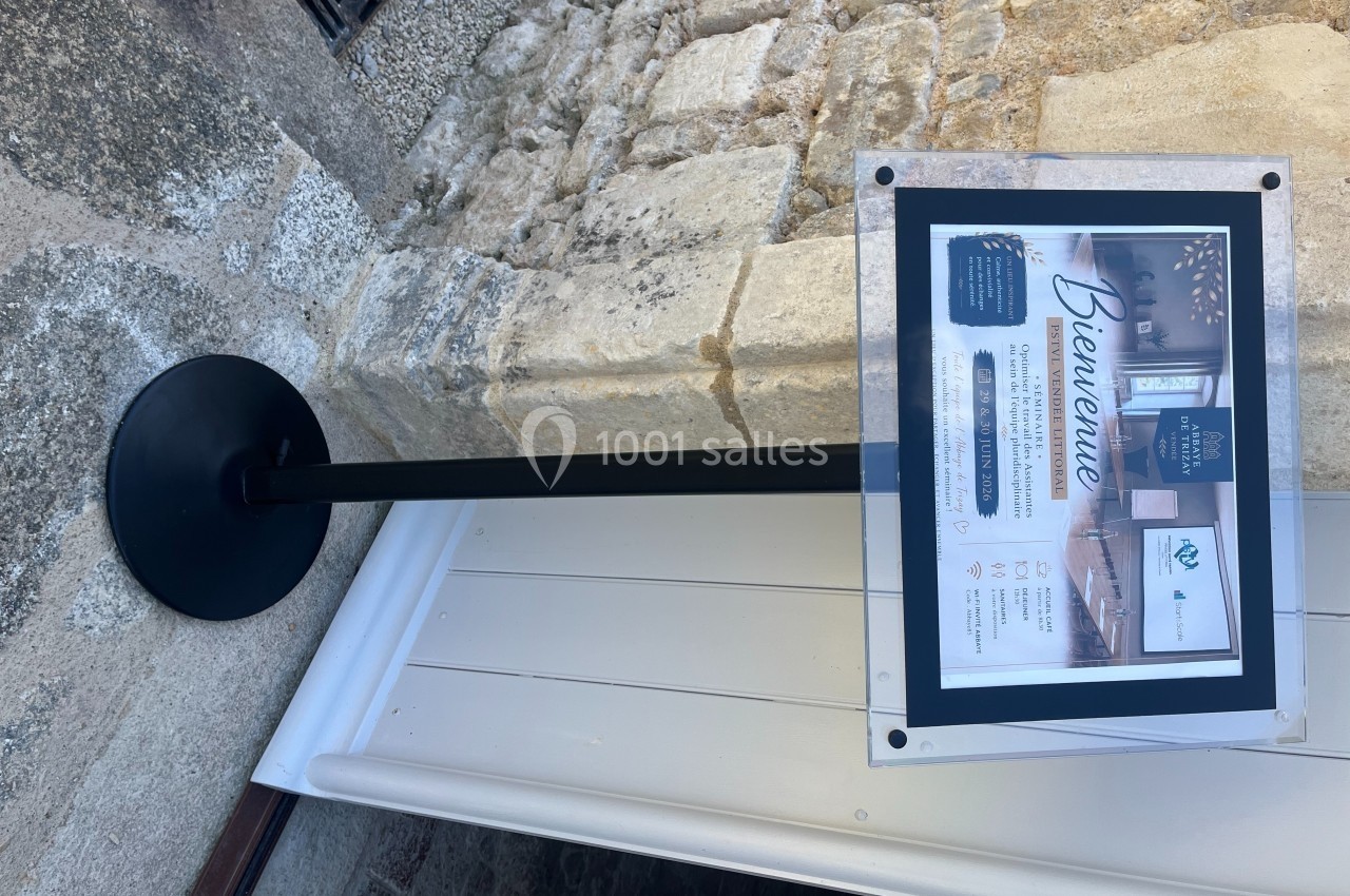 Panneau d'accueil sur pied indiquant des informations sur un lieu, fixé devant un mur en pierre.