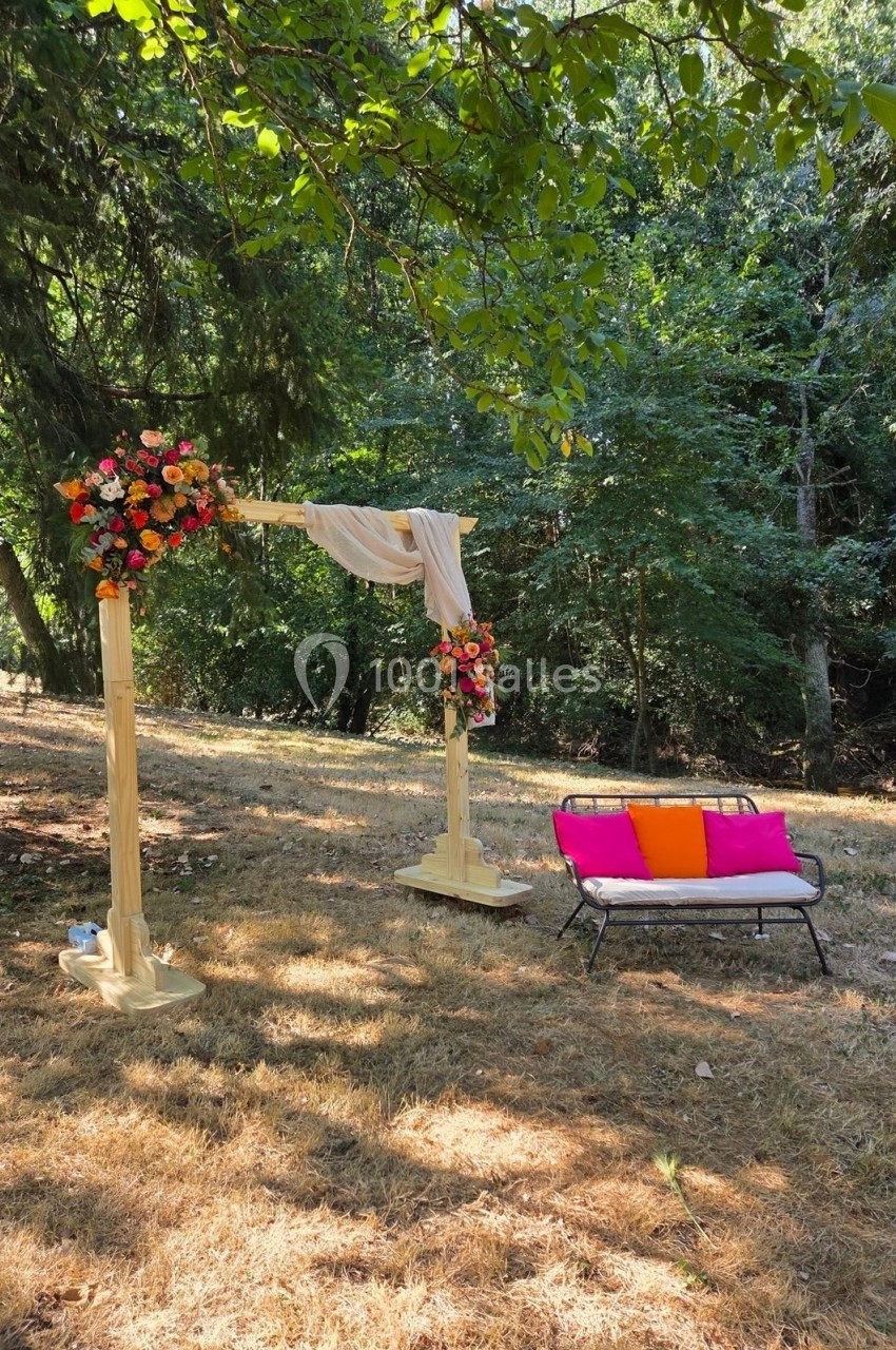 Arche en bois décorée de fleurs et voilage, avec un banc coloré, installés dans une clairière boisée.