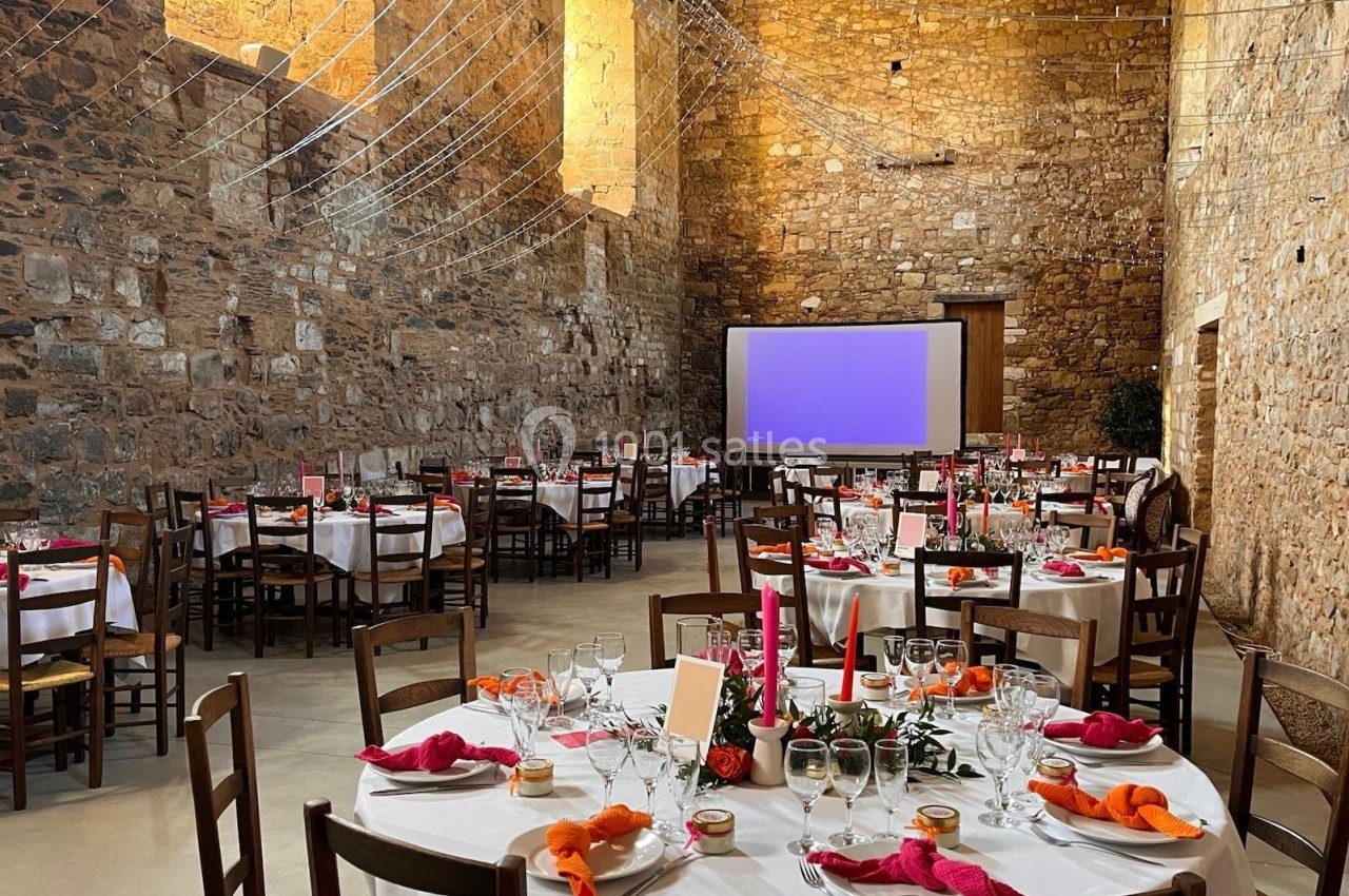 Salle en pierre aménagée pour un événement, avec tables rondes dressées, nappes blanches et décorations colorées.