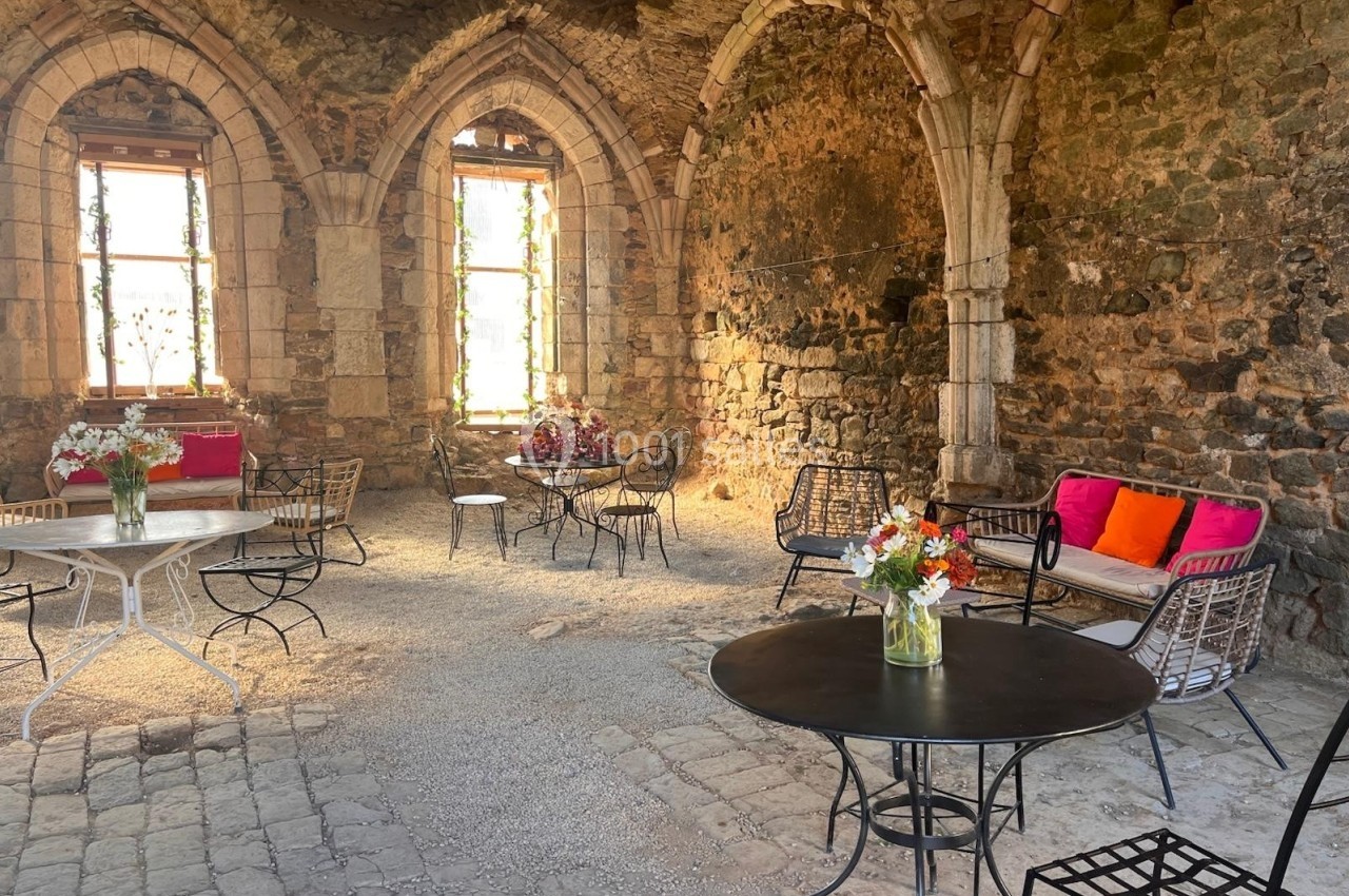Salle en pierre ancienne avec des tables, chaises et bouquets de fleurs, éclairée par des fenêtres à arc.