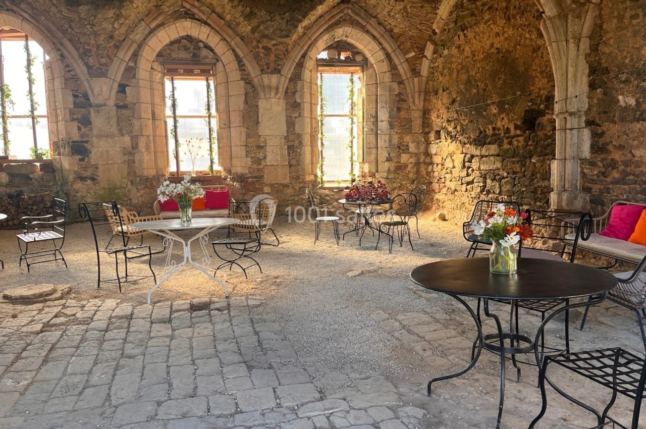Salle voûtée en pierre avec tables et chaises en fer forgé, décorée de fleurs et éclairée par des fenêtres cintrées.