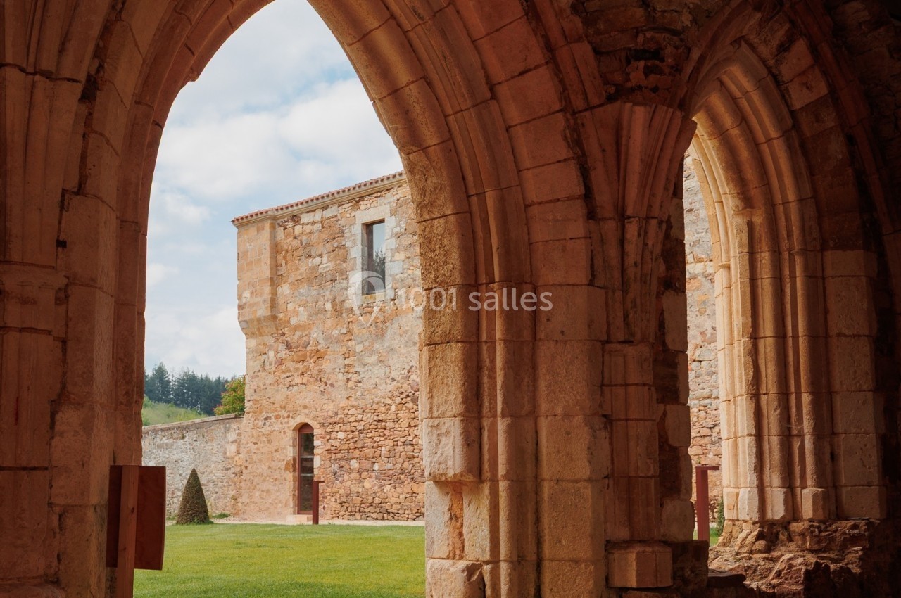 Vue depuis des arches en pierre donnant sur une cour verdoyante et un bâtiment ancien en pierre sous un ciel partiellement…