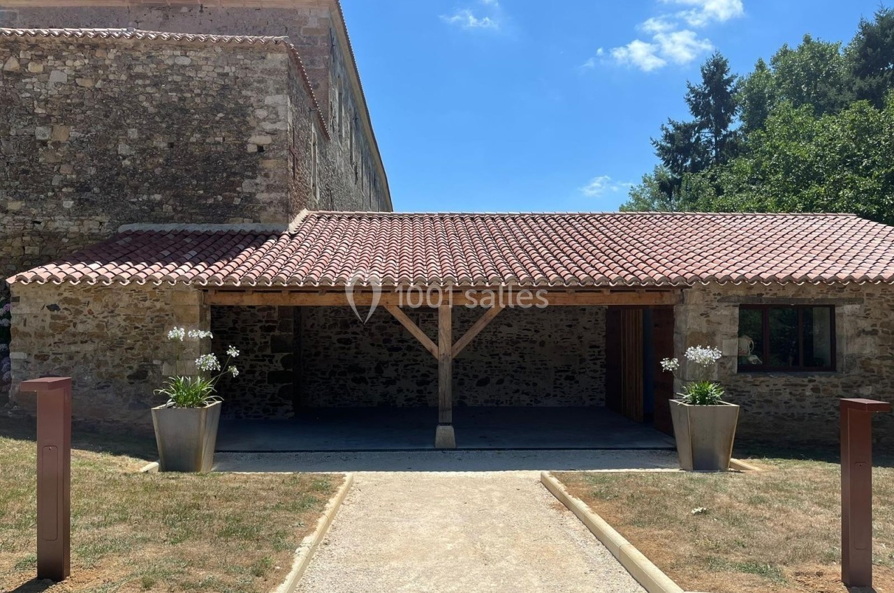 Façade d'un bâtiment en pierre avec un toit en tuiles, abri en bois et deux jardinières contenant des fleurs blanches.